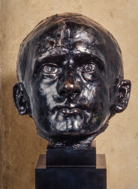A bronze sculpture of a man's head staring directly ahead. The facial features are detailed and precise. The man does not have any hair.