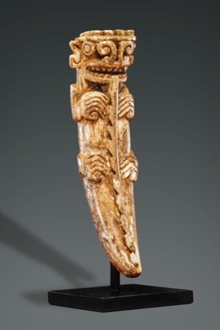 A crocodile pendant, tapering from the top to a pointed bottom with the head at the widest end, with large, coiled nostrils and a mouth full of teeth. The crocodile has hands and feet resting on the front on the pendant, pointing inward toward a vertical ridge with jagged edges, which runs down the center of the form.