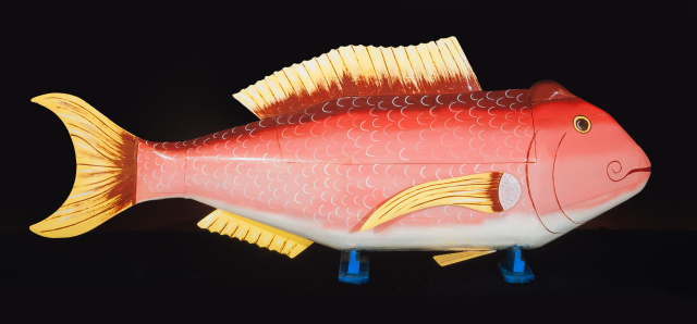 A painted wooden coffin in the shape of a fish. The fish is painted pink with red stripes near its top, and the fins are painted yellow, with crimson areas where the fins join with the fish's body.