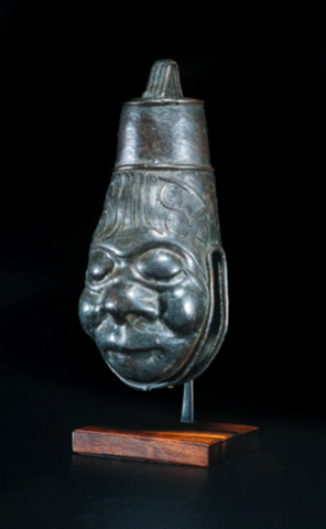 A bell in the form of a human head, with a handle at the top and an opening at the bottom, curving around the sides of the head and the chin. The head is wide and rounded at the bottom, then tapers upward to the top, wearing a cylindrical, tapering hat or headdress.