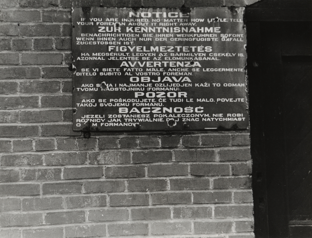 A horizontal black-and-white photograph depicts a close up of a metal sign posted on a brick wall. The sign reads, "NOTICE | IF YOU ARE INJURED, NO MATTER HOW LITTLE, TELL | YOUR FOREMAN ABOUT IT RIGHT AWAY." That phrase is then repeated in German, Hungarian, Italian, Lithuanian, Czech, and Polish.