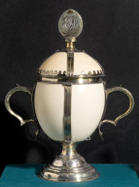 A white ostrich egg that has been transformed into a cup by adding a silver frame, base, handles, and an ornament on the lid. That ornament is oval-shaped and engraved with the monogram "EGJ."