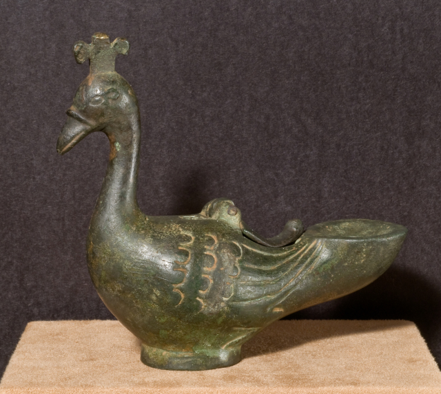 An oil lamp in the form of a peacock with a hinged cover over the oil reservoir on the back and a hole for the wick insertion on top of the truncated circular tail-end. There are incised details throughout the body.