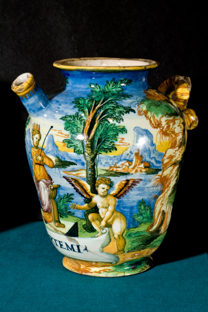 A small ceramic pot with a spout and a handle. It is decorated with a scene of a woman seated in a landscape, with angels holding a banner that reads: S. D. ARTEMI. The pot is decorated with blues, yellows, and greens.