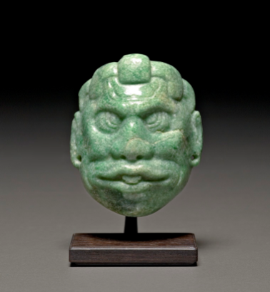 Light green jadeite pendant in the form of a head. The face has a wide nose, eyes with outlined pupils that point inward and upward, and a mouth that is slightly open, the tongue visible.